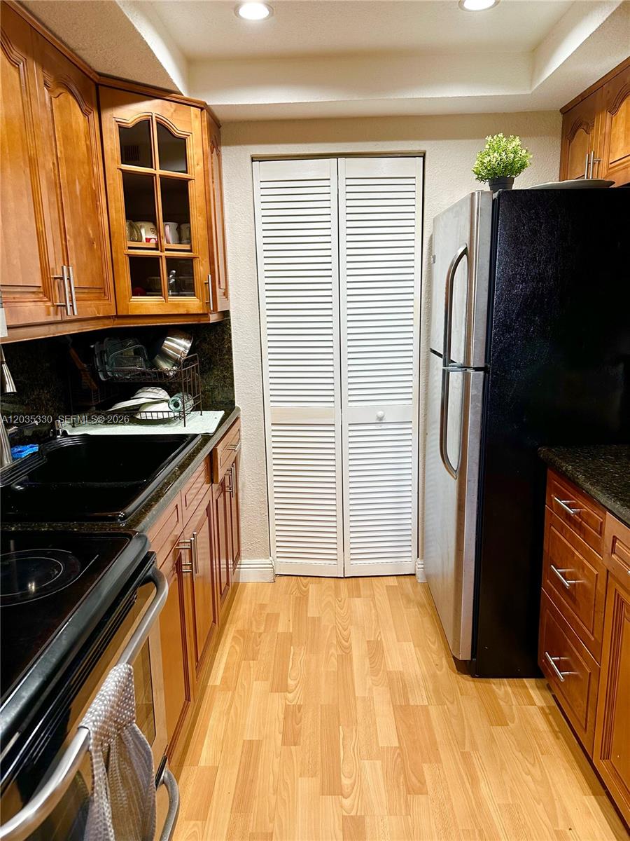 A galley kitchen features wood cabinets, dark countertops, a black refrigerator, and light wood flooring — 500 NE 2nd St # 327, Dania Beach FL 33004, DANIA BEACH, FL