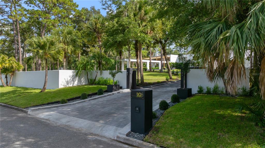 A paved driveway leads through a modern entrance gate toward a white house among trees — 808 SWEETWATER CLUB BOULEVARD, LONGWOOD, FL