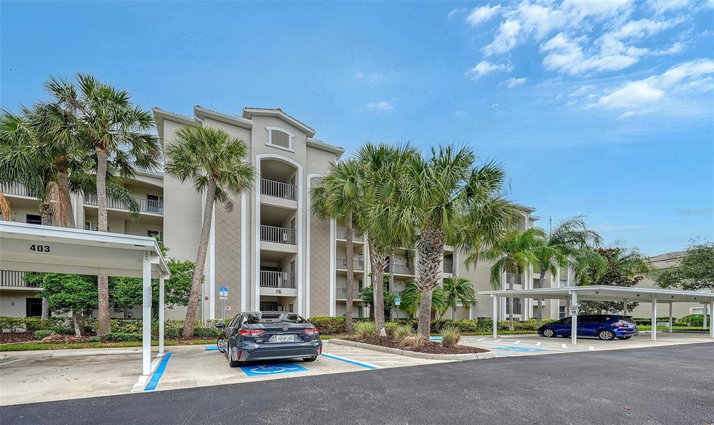 A multi-story apartment building with covered parking spaces and palm trees in the foreground — 7705 GRAND ESTUARY TRAIL UNIT 207, BRADENTON, FL