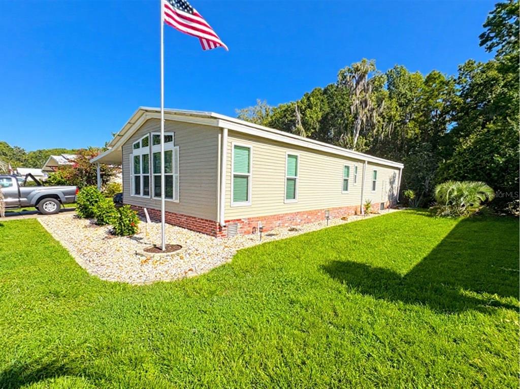 A tan manufactured home with a brick base, gravel landscaping, and an American flag pole — 601 ROBIN LANE, WILDWOOD, FL