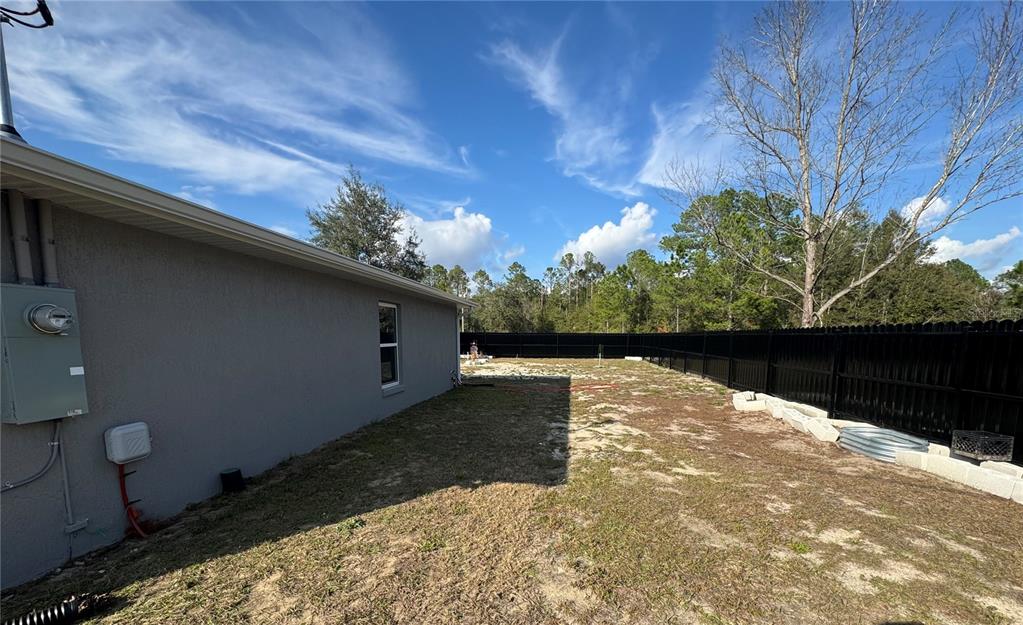 The side yard of a house features a gray stucco wall, a black fence, and sandy ground — 13183 SW 113TH PLACE, DUNNELLON, FL