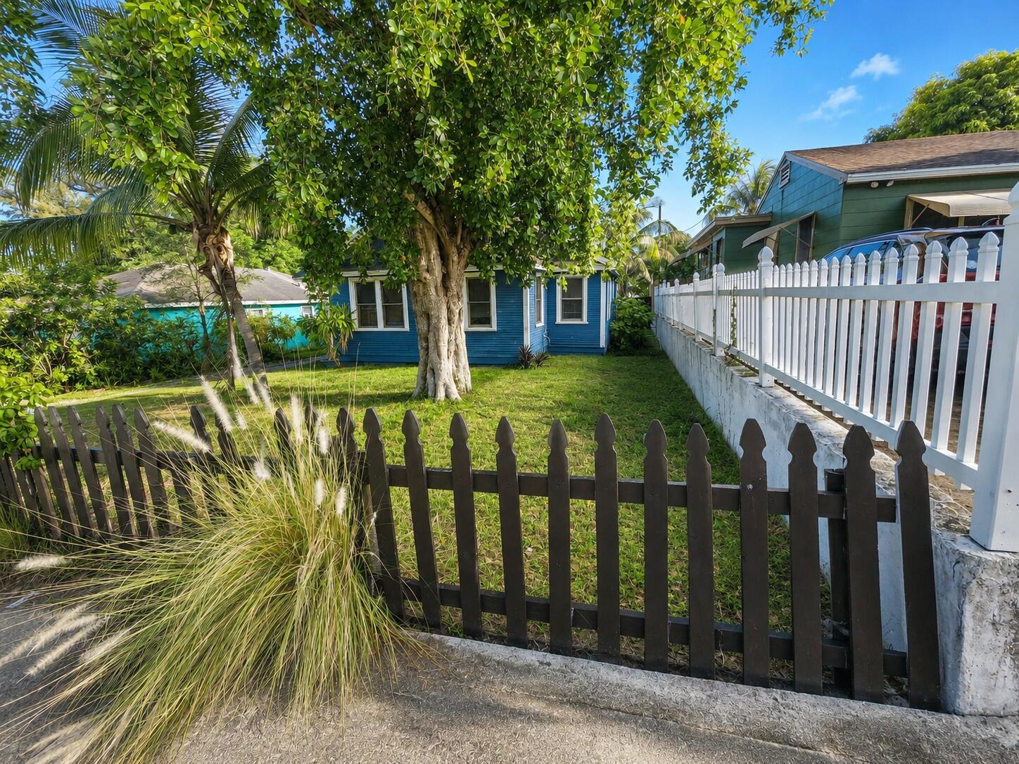 A blue house sits behind a dark wooden fence and a large tree in a yard — 1736 7th Avenue N, FL 33460, FL