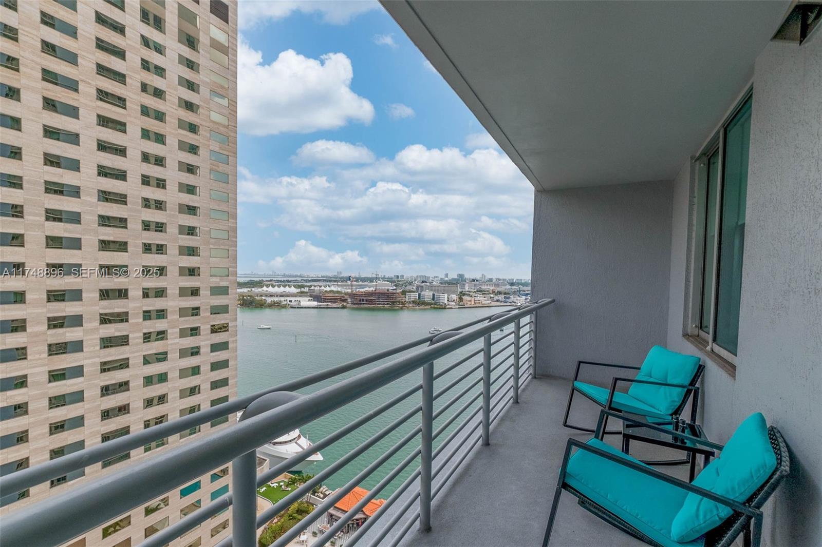 A balcony with two turquoise chairs overlooks a body of water and a tall building — 335 S Biscayne Blvd # 2402, Miami FL 33131, MIAMI, FL