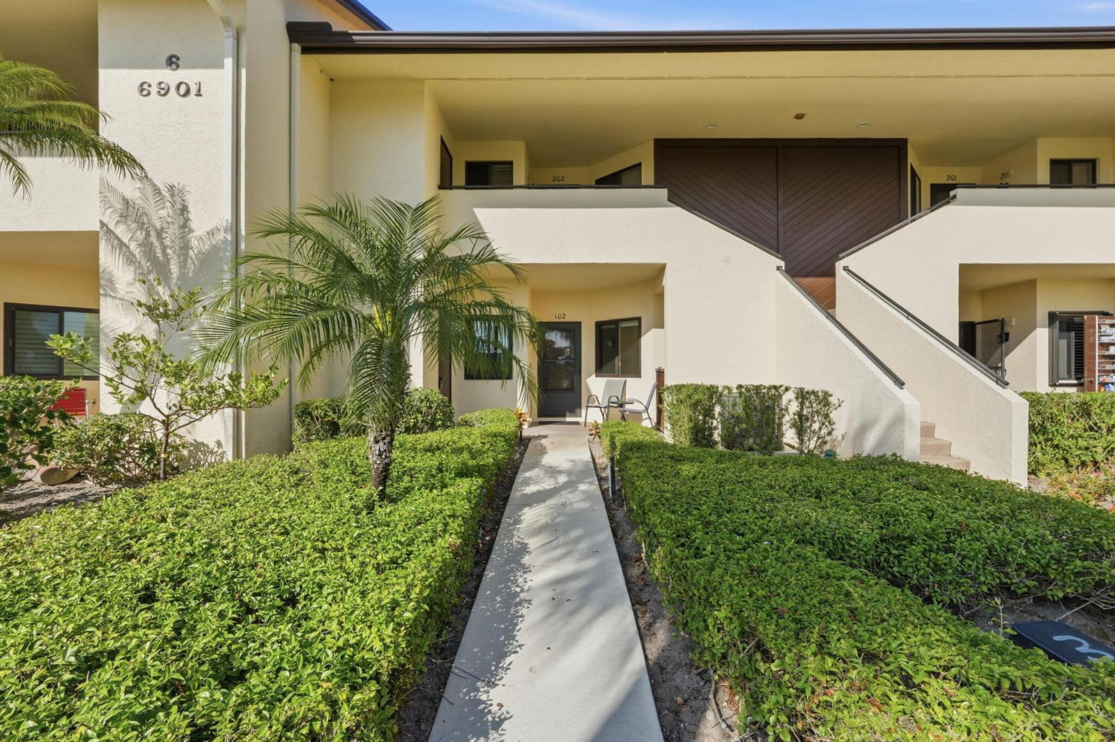 A two-story apartment building exterior features a concrete walkway leading to a ground-floor entrance — 6901 SE Constitution Boulevard # 102, Hobe Sound FL 33455, HOBE SOUND, FL