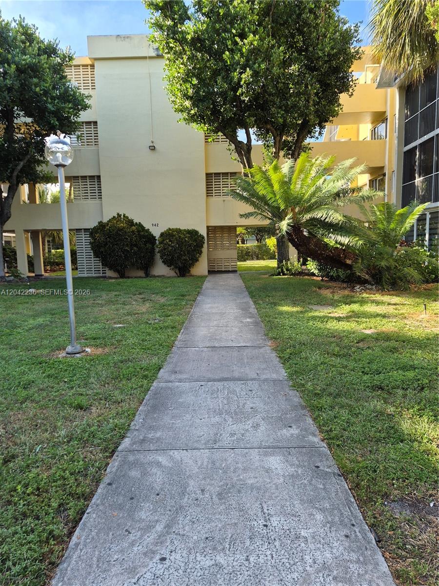 A concrete walkway leads to the entrance of a multi-story yellow apartment building — 942 NE 199th St # 4G, Miami FL 33179, MIAMI, FL