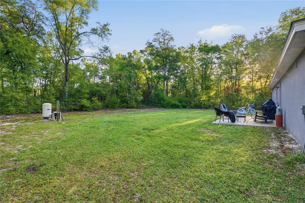 A grassy backyard features a concrete patio with outdoor furniture and a wooded backdrop — 15541 NE 13TH LANE, WILLISTON, FL