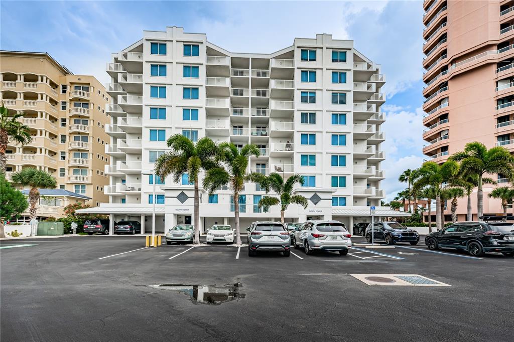 A multi-story white residential building stands behind a parking lot with several parked cars — 1350 GULF BOULEVARD UNIT 703, CLEARWATER BEACH, FL