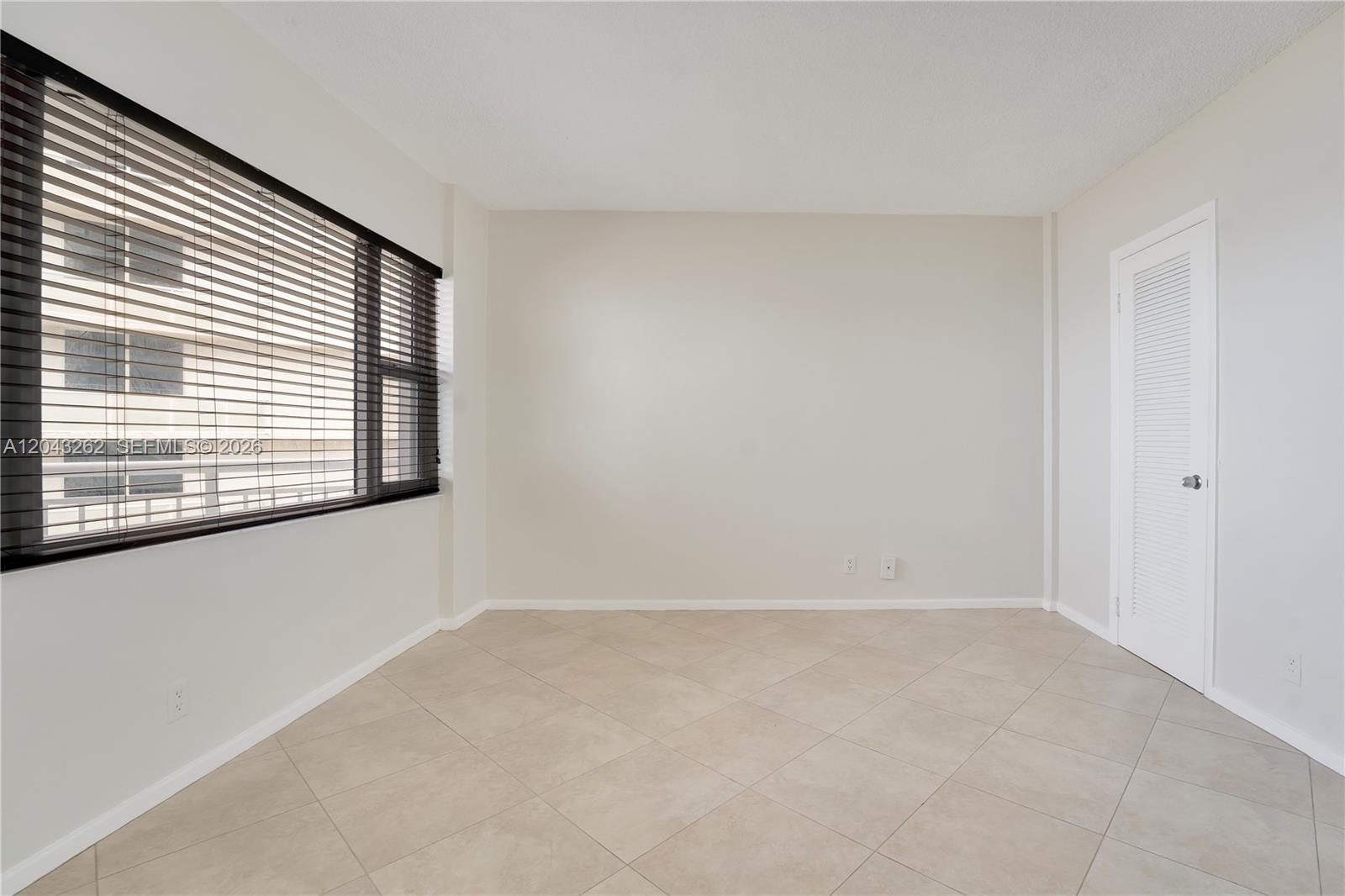 An empty room features beige tile flooring, white walls, a large window with blinds, and a door — 710 N Ocean Blvd # 710, Pompano Beach FL 33062, POMPANO BEACH, FL