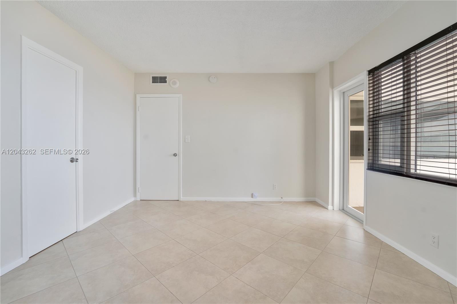 An empty room features beige tile flooring, white walls, two doors, and a window with blinds — 710 N Ocean Blvd # 710, Pompano Beach FL 33062, POMPANO BEACH, FL