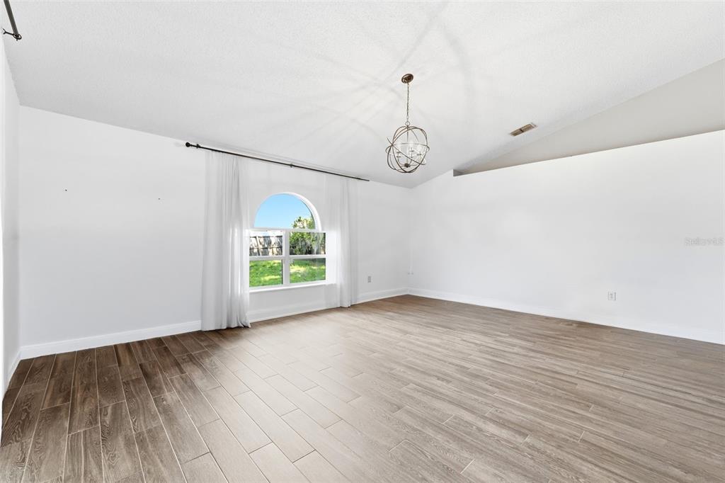 An empty room features wood-look tile flooring, white walls, and a vaulted ceiling with a chandelier — 1176 LYRIC DRIVE, DELTONA, FL
