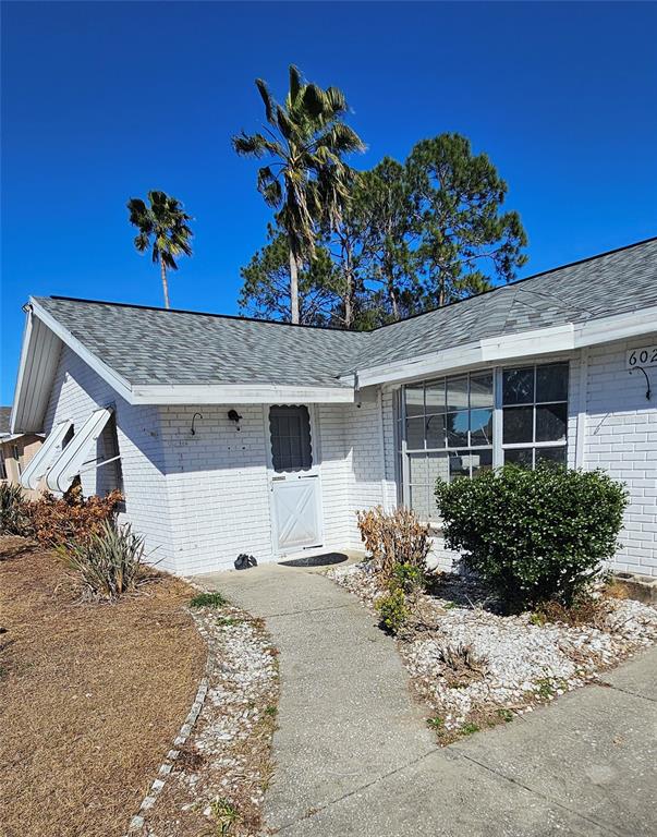 A white brick house features a gray shingled roof, a front walkway, and palm trees — 6020 17TH STREET, ZEPHYRHILLS, FL