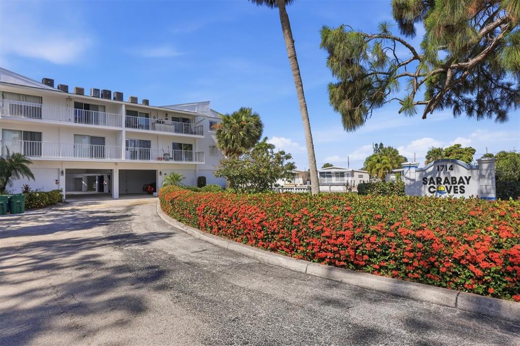 A three-story apartment building stands behind a curved hedge of red flowers — 1714 69TH AVENUE W UNIT A303, BRADENTON, FL