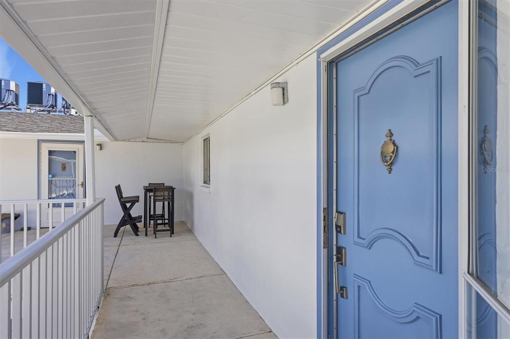 A covered outdoor walkway features a blue front door and a small table with chairs — 1714 69TH AVENUE W UNIT A303, BRADENTON, FL
