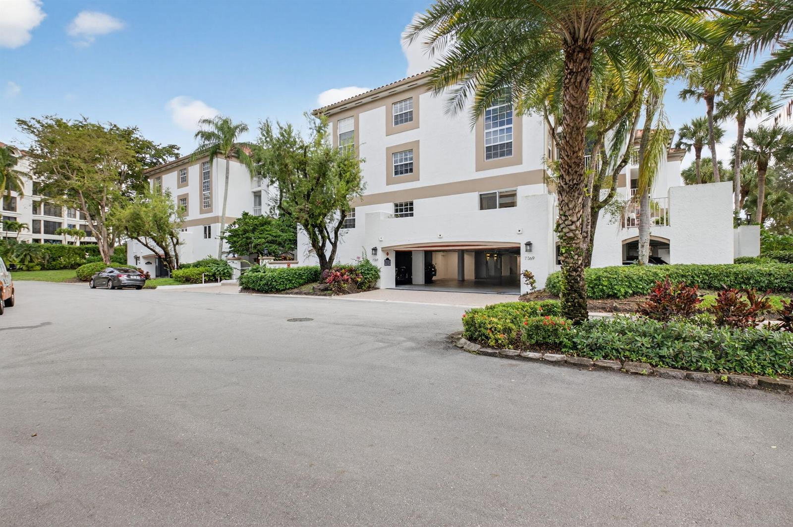 A multi-story white residential building features a ground-level parking garage and surrounding tropical landscaping — 7369 Orangewood Lane # 307, Boca Raton FL 33433, BOCA RATON, FL