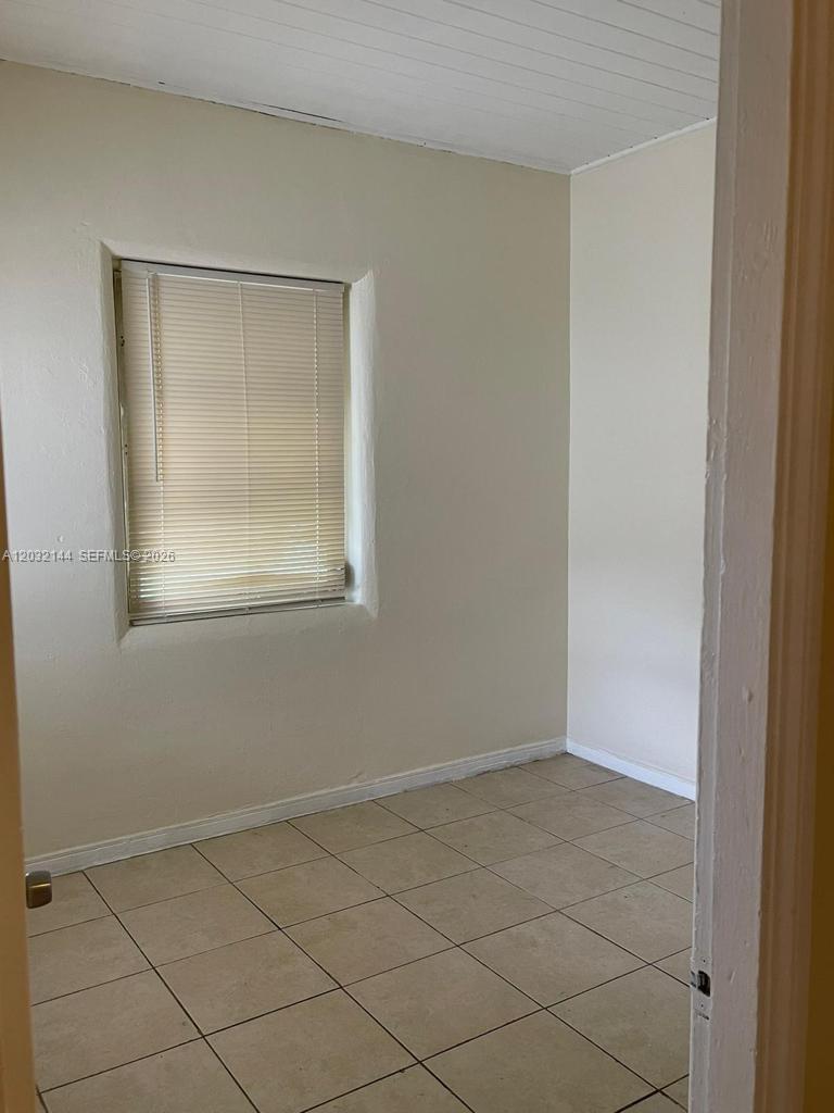 A small, empty room features light-colored walls, tiled flooring, and a single window with blinds — MIAMI, FL