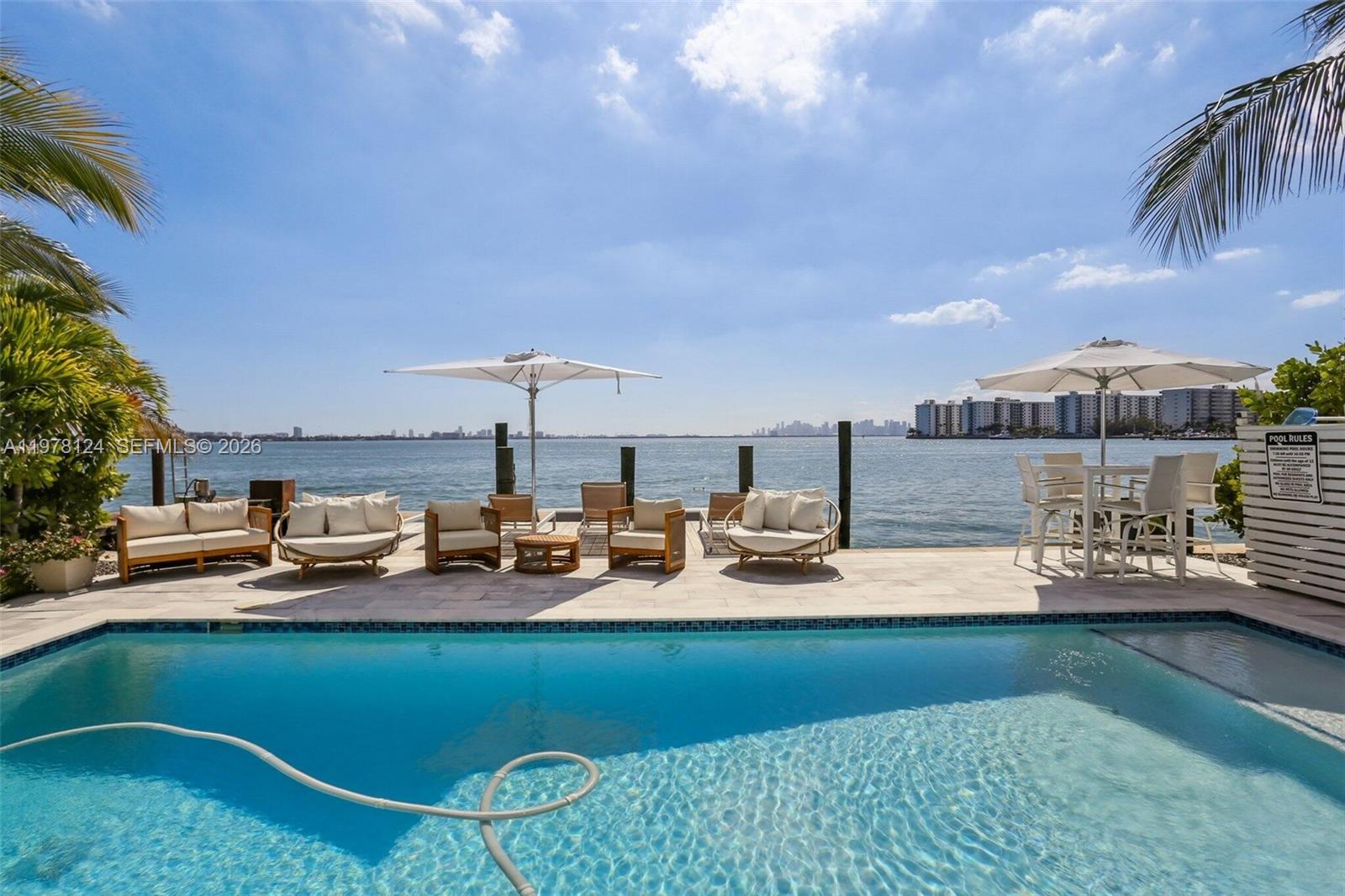 A swimming pool overlooks a bay with outdoor seating, umbrellas, and a distant city skyline — 1930 Bay Dr # 4, Miami Beach FL 33141, MIAMI BEACH, FL