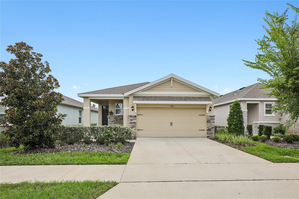 A tan single-story house with a two-car garage and stone accents sits behind a driveway — 5395 SUNSHINE DRIVE, WILDWOOD, FL