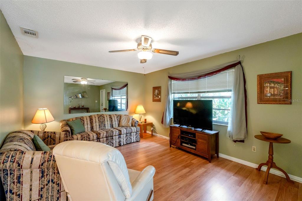 A living room features a patterned sofa, a white recliner, a television, and wood flooring — 1814 FOXHUNT DRIVE UNIT A, SUN CITY CENTER, FL