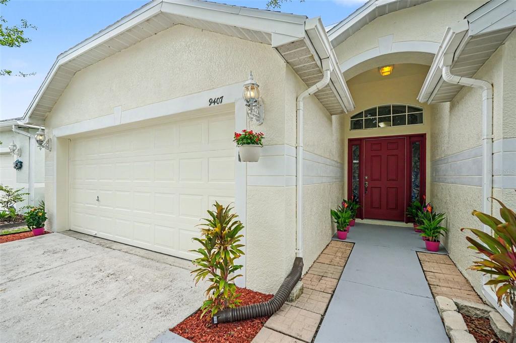A beige house exterior features a white garage door, a red front door, and landscaping — 9407 CYPRESS HARBOR DRIVE, GIBSONTON, FL