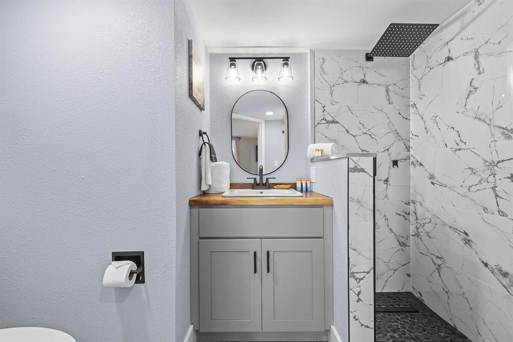A bathroom features a gray vanity with a wood countertop, oval mirror, and marble-tiled shower — 421 S ATLANTIC AVENUE UNIT 103, NEW SMYRNA BEACH, FL