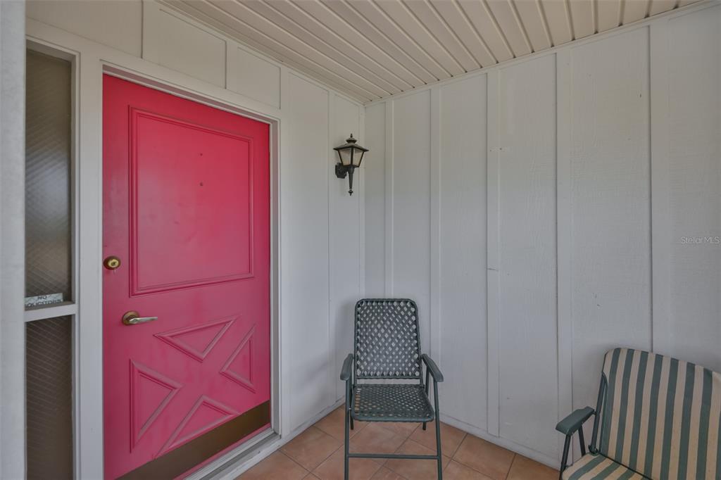A covered porch features a bright pink front door, tiled flooring, and two outdoor chairs — 1525 ALLEGHENY DRIVE, SUN CITY CENTER, FL