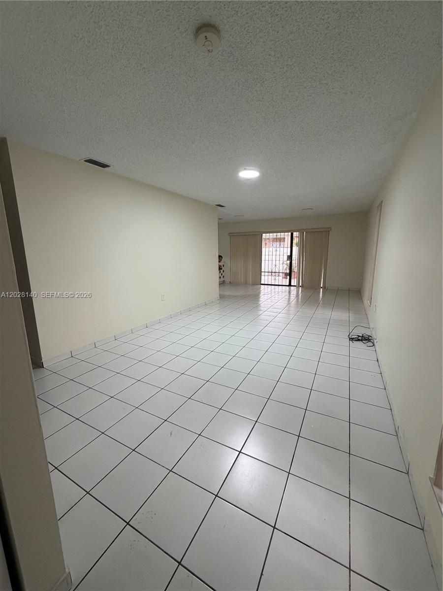 A long, empty living room features white tile flooring, plain walls, and a sliding glass door — MIAMI, FL