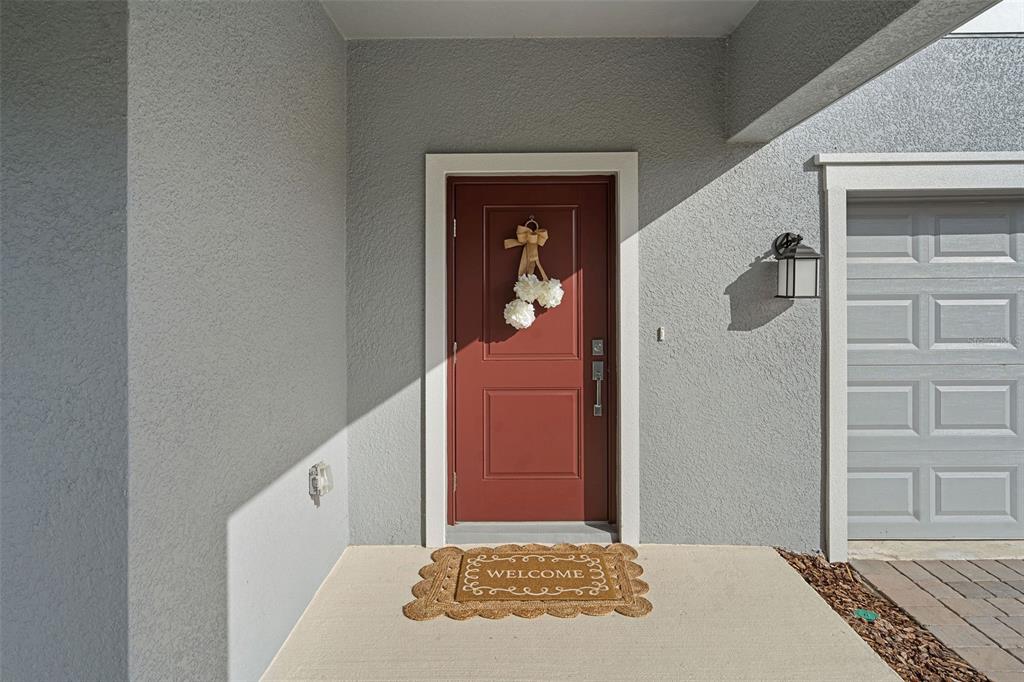 A red front door with a wreath and welcome mat sits under a covered entryway — 3000 NAMASTE DRIVE, CLERMONT, FL