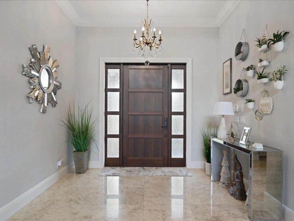 A bright entryway features a dark wood front door, a chandelier, and a mirrored console table — 10596 Versailles Boulevard, Wellington FL 33449, WELLINGTON, FL