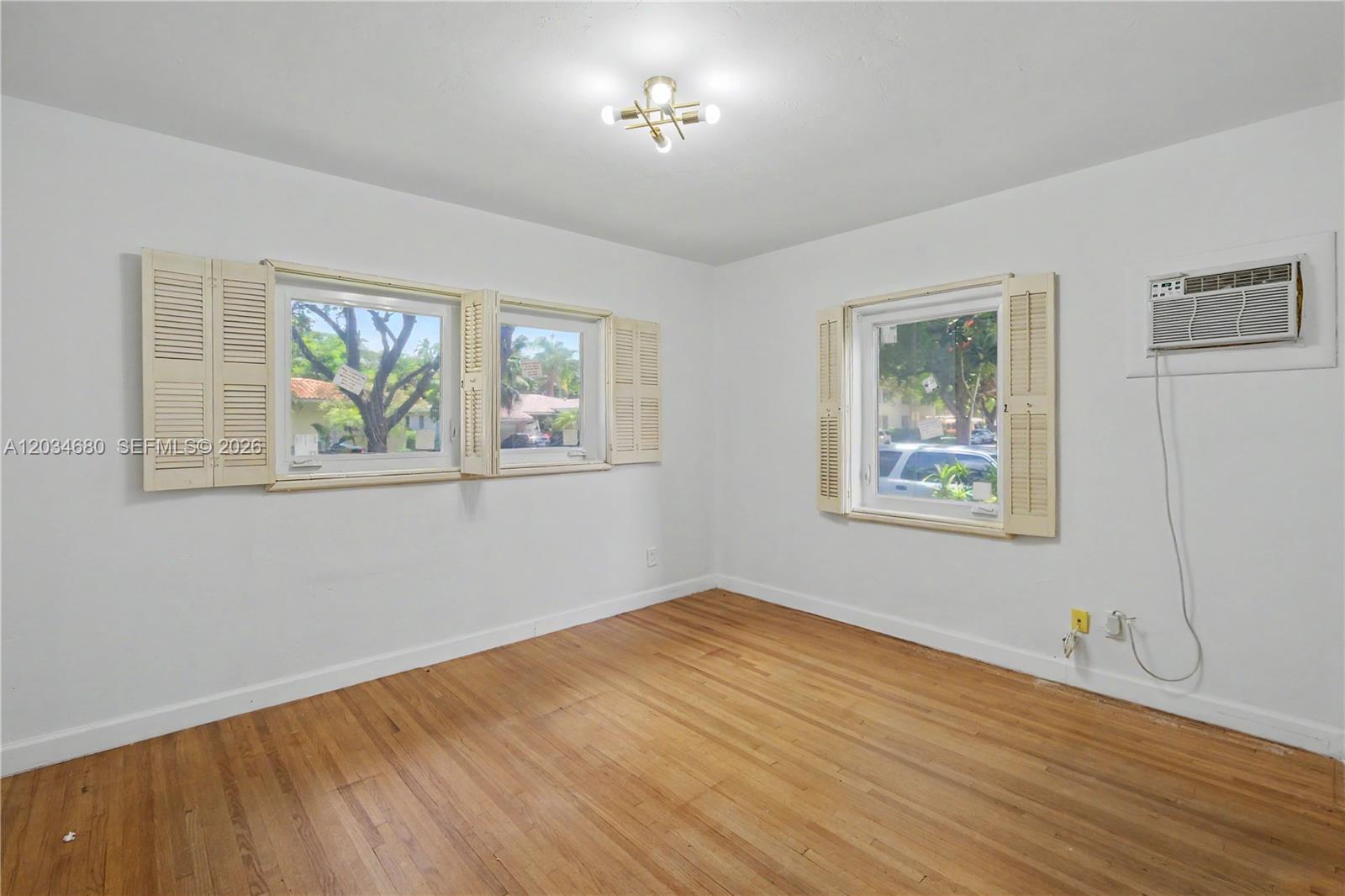 An empty room features white walls, hardwood floors, three windows with shutters, and an AC unit — 236 Candia Ave, Coral Gables FL 33134, CORAL GABLES, FL