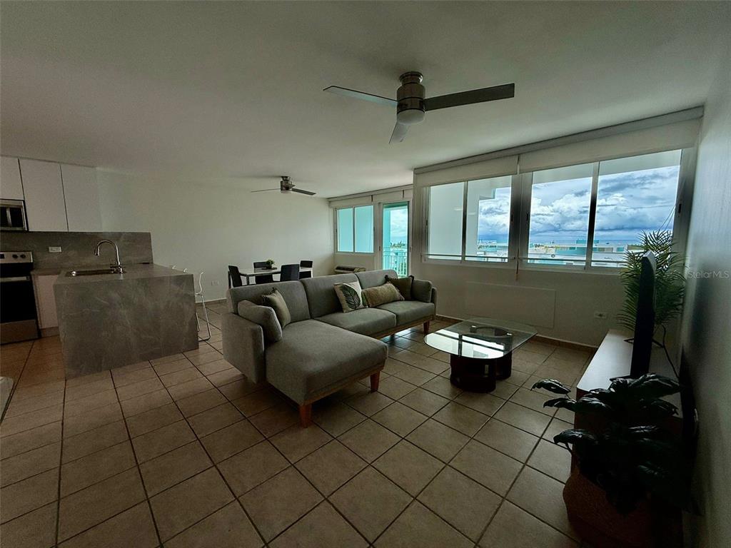 A living room with a gray sectional sofa, tile floors, and large windows overlooking the ocean — 96QJ+3RG CLL LAS COLINAS UNIT 301, RIO GRANDE, PR