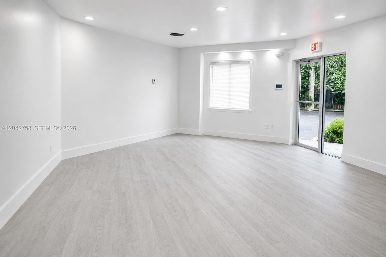 An empty room features light wood-look flooring, white walls, recessed lighting, and a glass exit door — 9450 NW 58th St, Doral FL 33178, DORAL, FL