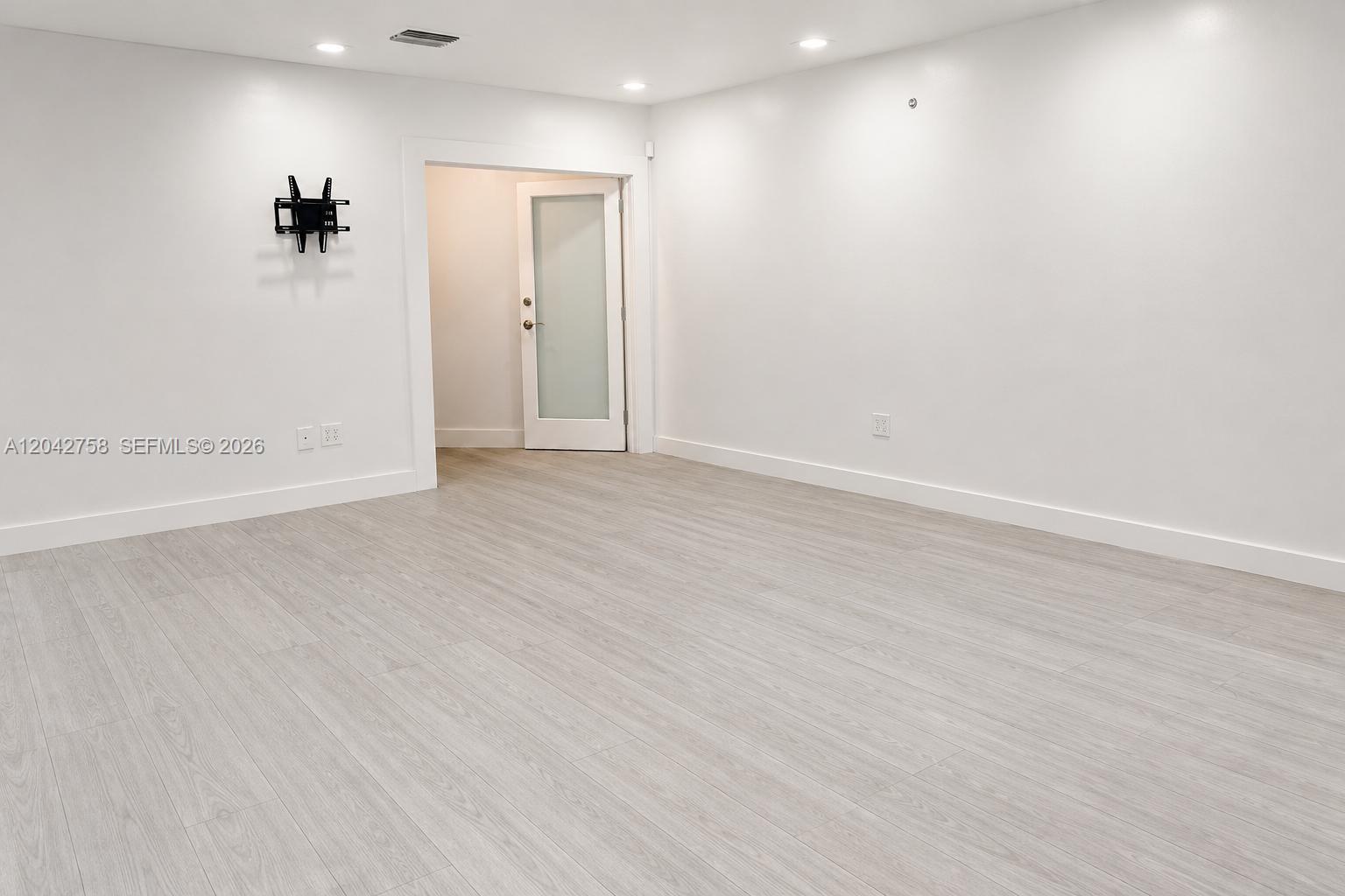 An empty room features light wood-look flooring, white walls, recessed lighting, and a wall-mounted TV bracket — 9450 NW 58th St, Doral FL 33178, DORAL, FL