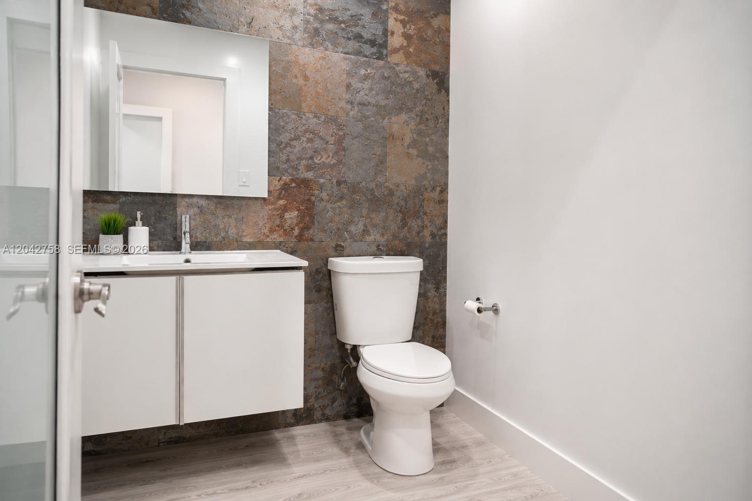 A bathroom features a white vanity, a toilet, and a wall with rustic stone tiles — 9450 NW 58th St, Doral FL 33178, DORAL, FL