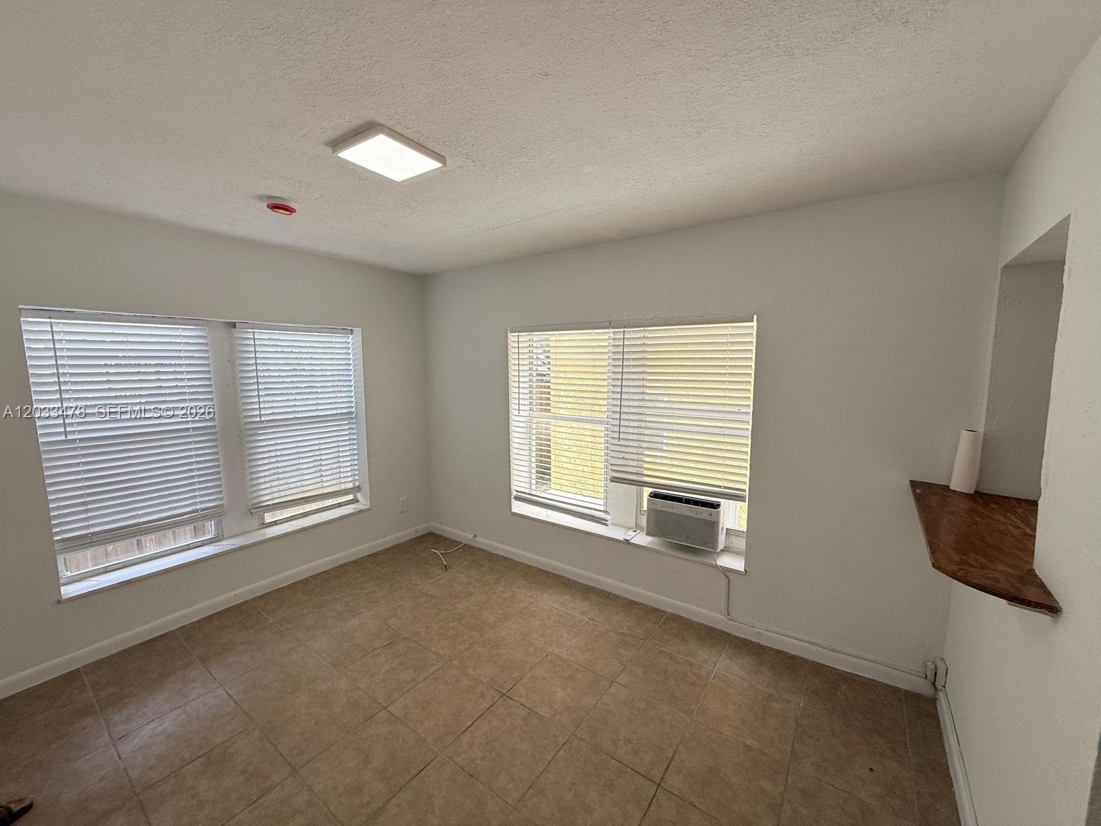 An empty room features tile flooring, white walls, two windows with blinds, and an air conditioner — 1612 Harrison St # 5, Hollywood FL 33020, HOLLYWOOD, FL