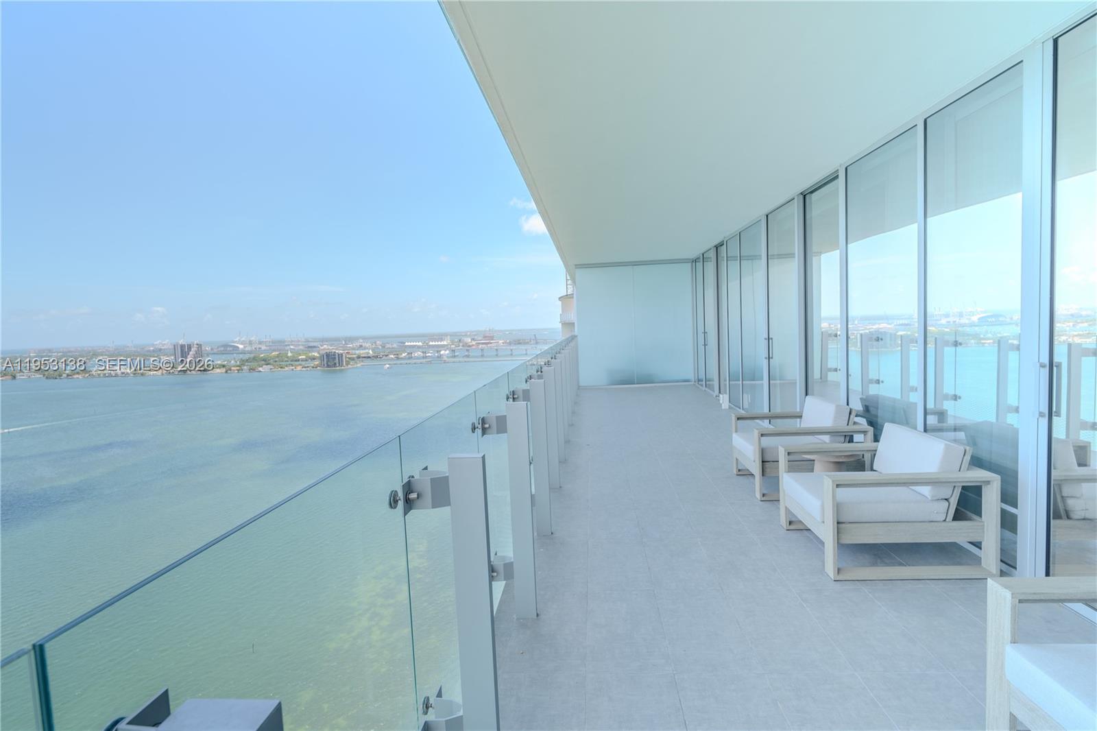 A high-rise balcony features a glass railing, light-colored tile flooring, and outdoor seating overlooking a bay — 700 NE 26th Ter # 2602, Miami FL 33137, MIAMI, FL