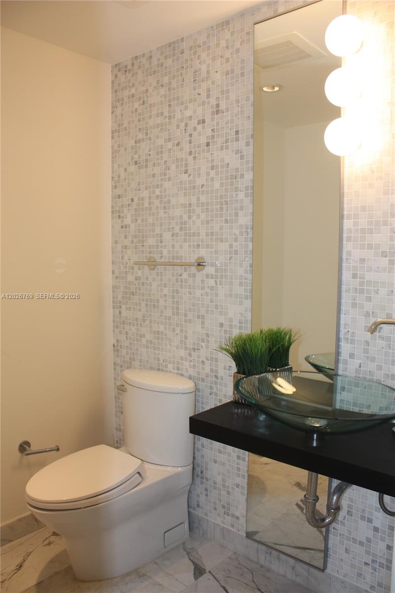 A bathroom features a glass vessel sink, a white toilet, and marble mosaic tile walls — 200 Biscayne Boulevard Way # 4211, Miami FL 33131, MIAMI, FL