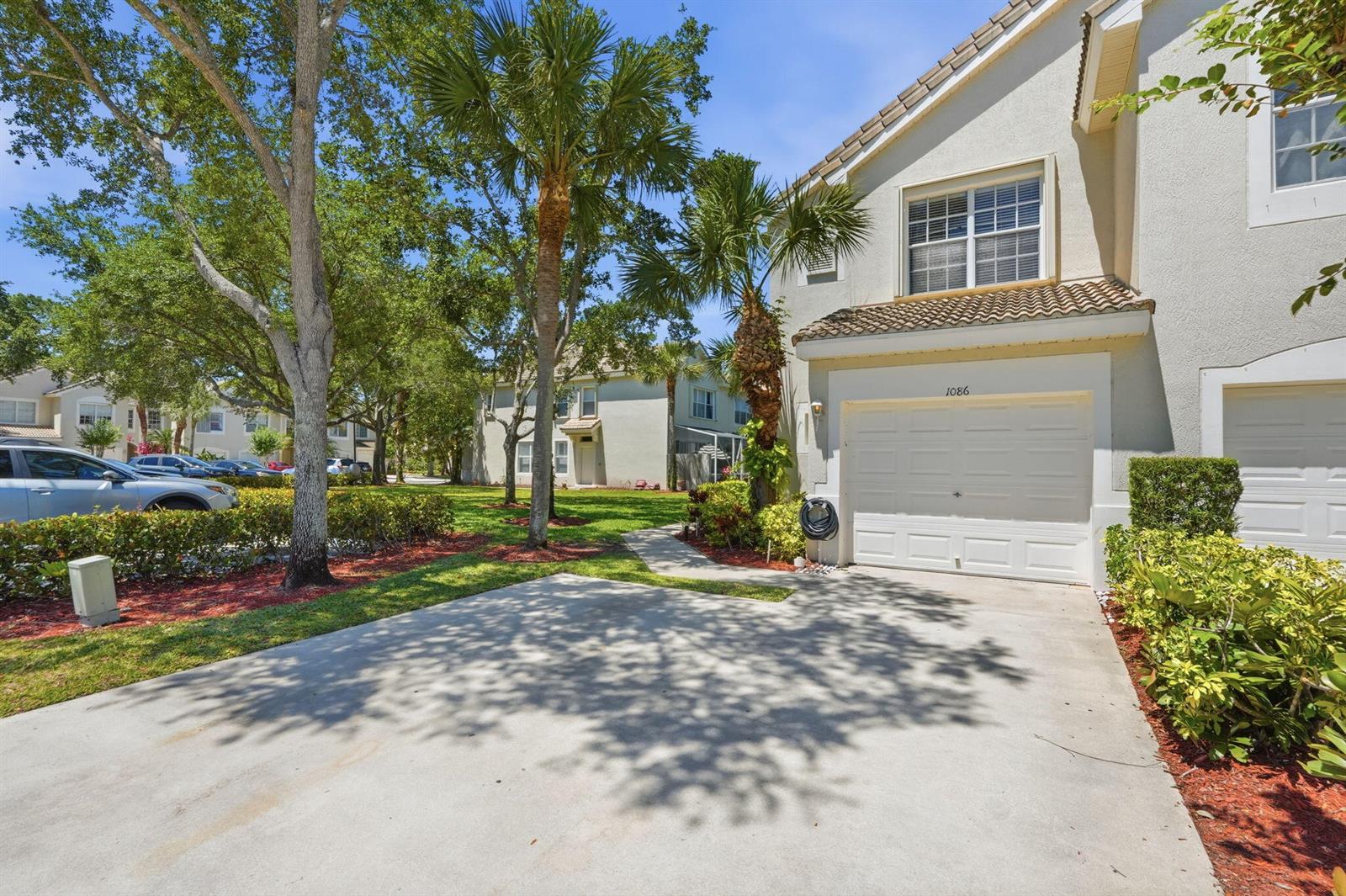 A two-story townhouse exterior features a concrete driveway, a white garage door, and palm trees — 1086 Woodfield Road, FL 33415, FL