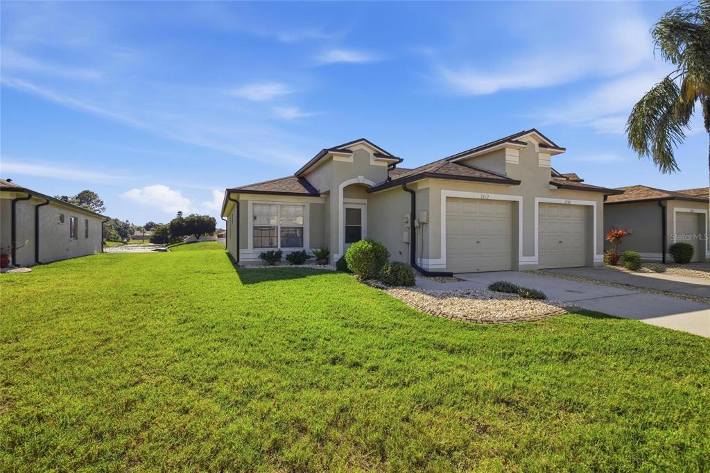 A single-story duplex with a tan stucco exterior, two garage doors, and a grassy lawn — 13307 ASHBARK COURT, RIVERVIEW, FL