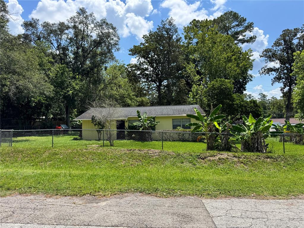 A yellow single-story house sits behind a chain-link fence surrounded by trees and plants — 921 NW 55TH TERRACE, GAINESVILLE, FL