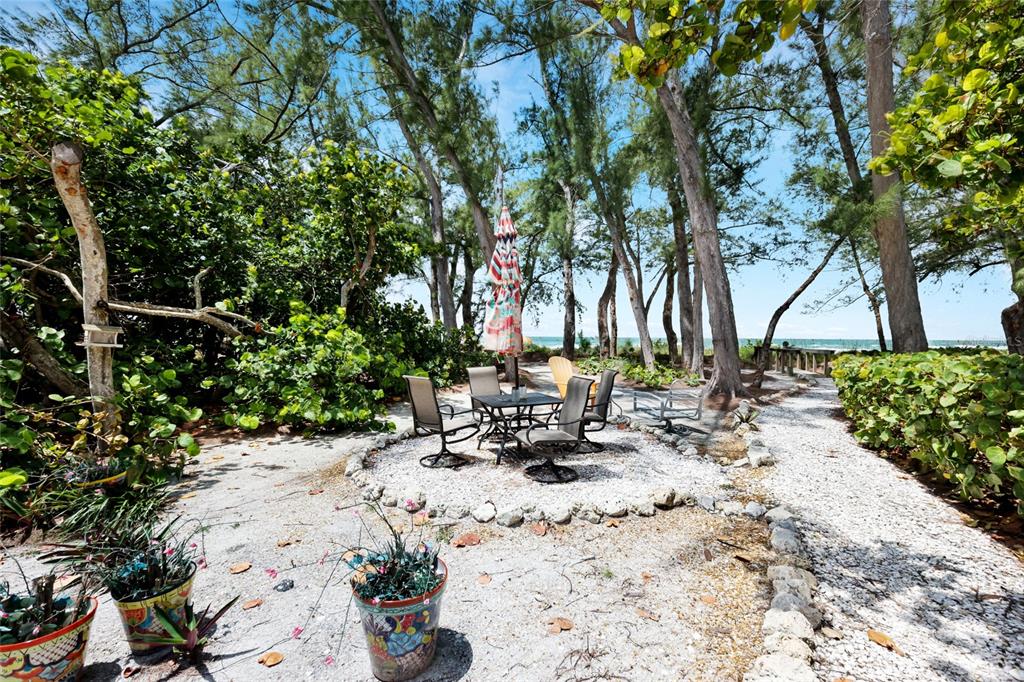 A gravel patio with a dining set and umbrella sits among trees near the beach — 103 51ST STREET, BRADENTON BEACH, FL