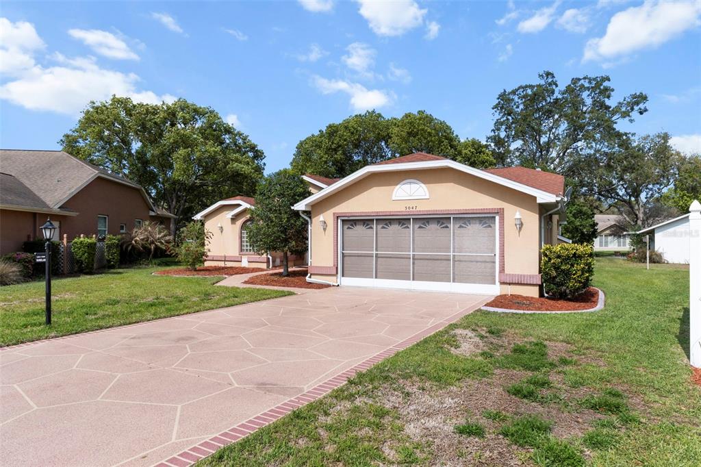 A tan single-story house features a screened garage door and a patterned concrete driveway — 3047 RIPPLING BROOK WAY, SPRING HILL, FL