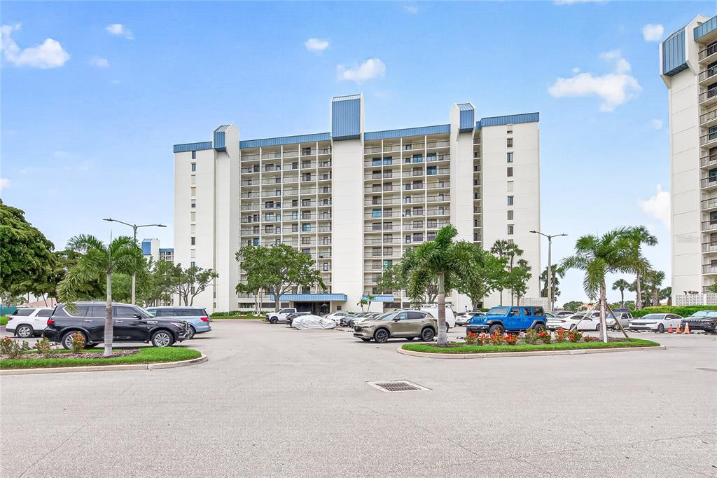 A multi-story residential building with balconies stands behind a paved parking lot with cars — 9425 BLIND PASS ROAD UNIT 1103, ST PETE BEACH, FL
