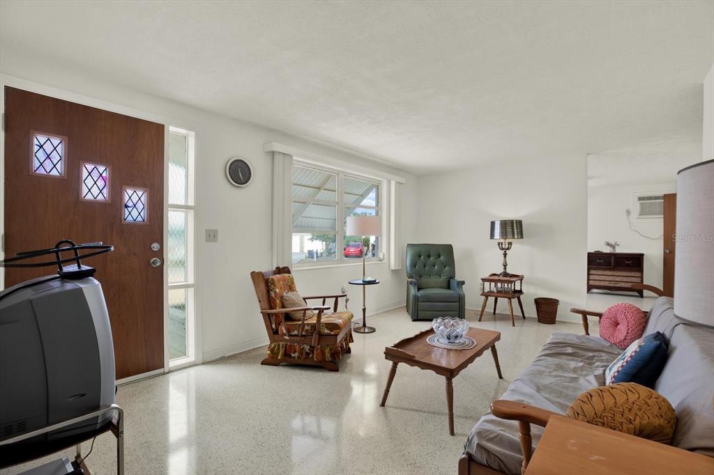 A living room features a wooden front door, terrazzo flooring, and a mix of vintage furniture — 1617 DERRICK STREET, HOLIDAY, FL