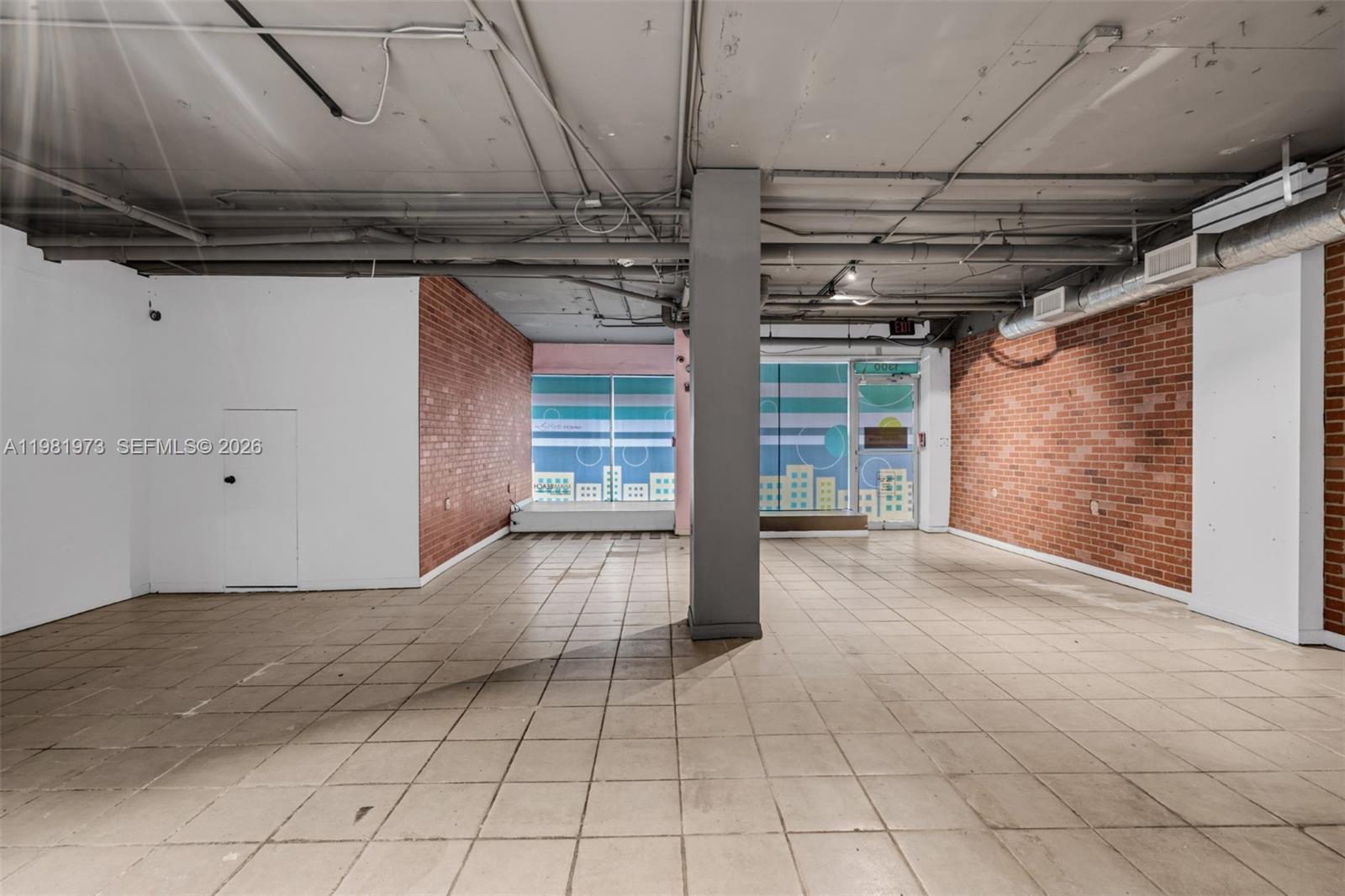 An empty commercial space features exposed ceiling pipes, brick walls, and tiled flooring — 1300 Lincoln Rd # 1B, Miami Beach FL 33139, MIAMI BEACH, FL