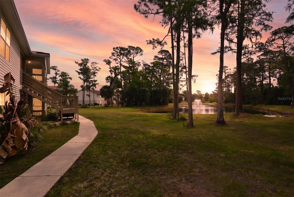 A concrete walkway leads past a multi-story building toward a pond with a fountain at sunset — 1600 BIG TREE ROAD UNIT G3, SOUTH DAYTONA, FL