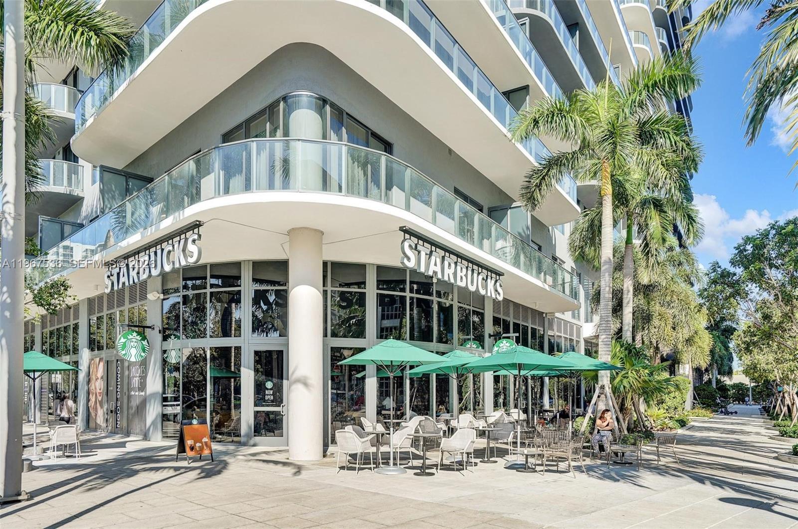 A modern corner building features a street-level Starbucks with outdoor patio seating under green umbrellas — 121 NE 34th St # 1705, Miami FL 33137, MIAMI, FL