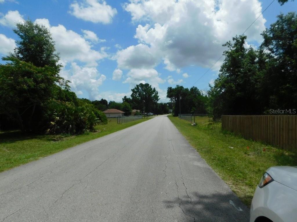 A paved road stretches forward between grassy shoulders, trees, and residential fencing under a cloudy sky — 0 NW 66TH STREET, OCALA, FL