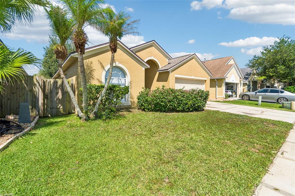 A single-story tan house with a two-car garage, palm trees, and a front lawn — 1229 CRIMSON CLOVER LANE, WESLEY CHAPEL, FL
