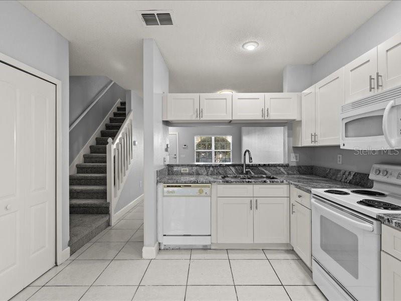 A kitchen with white cabinets, granite countertops, and tile flooring next to a carpeted staircase — 29924 PLAYA DEL REY LANE, WESLEY CHAPEL, FL