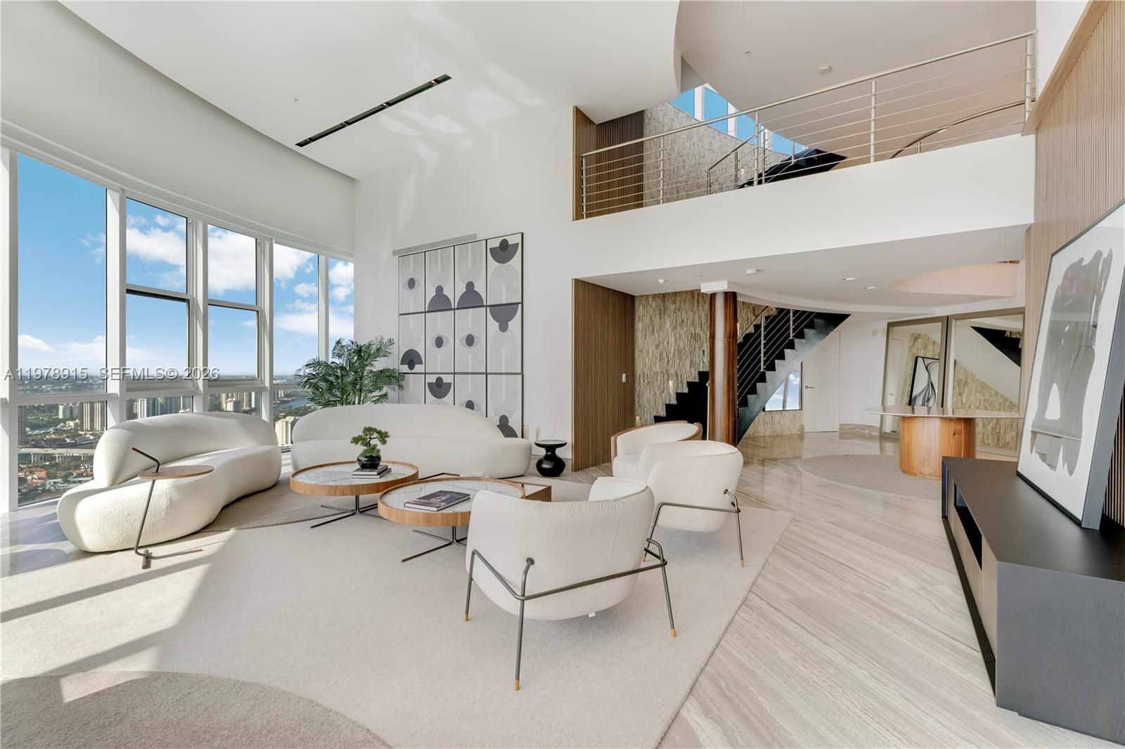 A modern, high-ceilinged living room features white curved sofas, a staircase, and floor-to-ceiling windows — 18201 E Collins Ave, Sunny Isles Beach FL 33160, SUNNY ISLES BEACH, FL